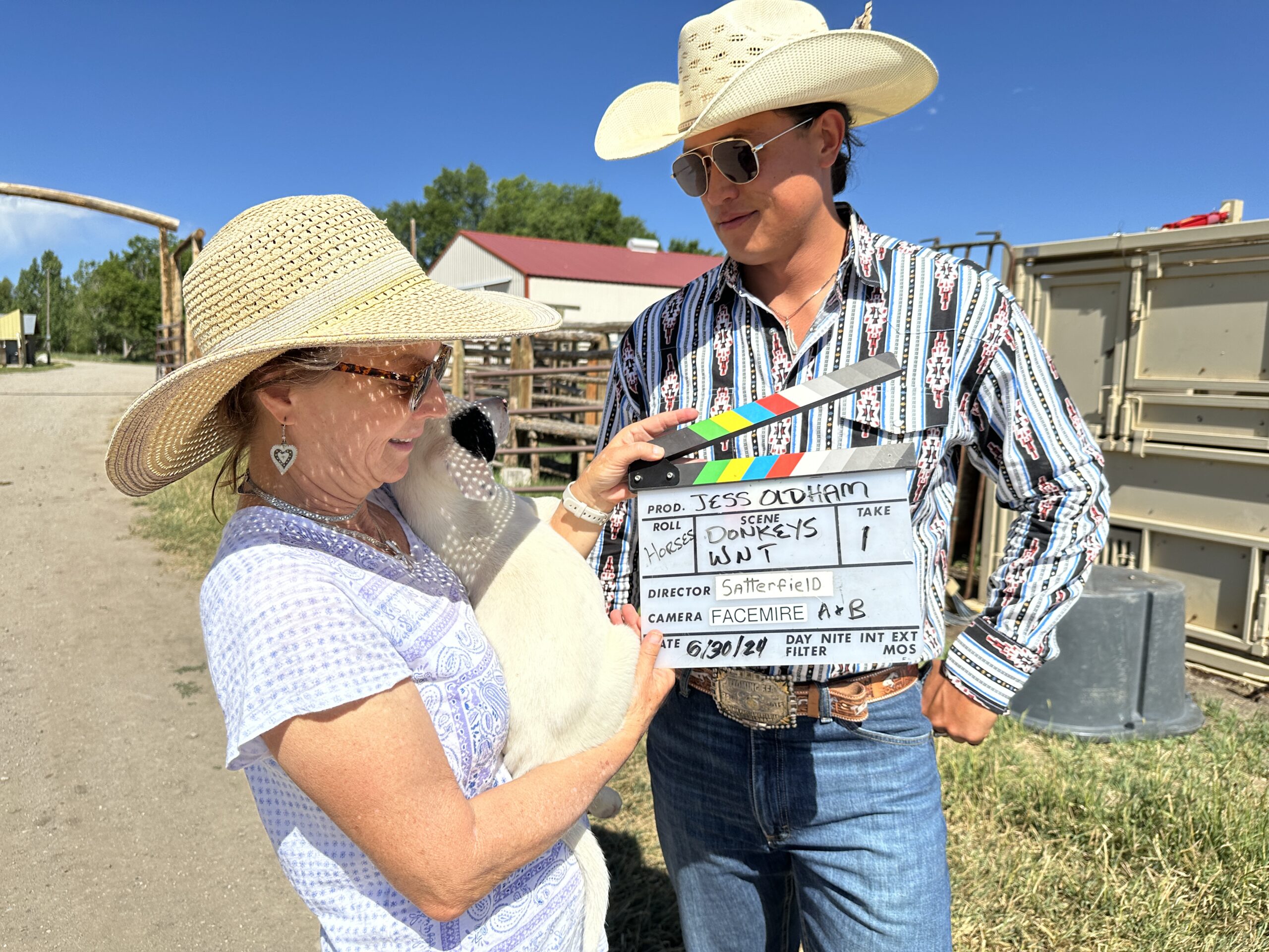 A Man and Woman in Cowboy Hats Hold a Movie Clapperboard, Smiling at the Camera in a Rustic Outdoor Setting.